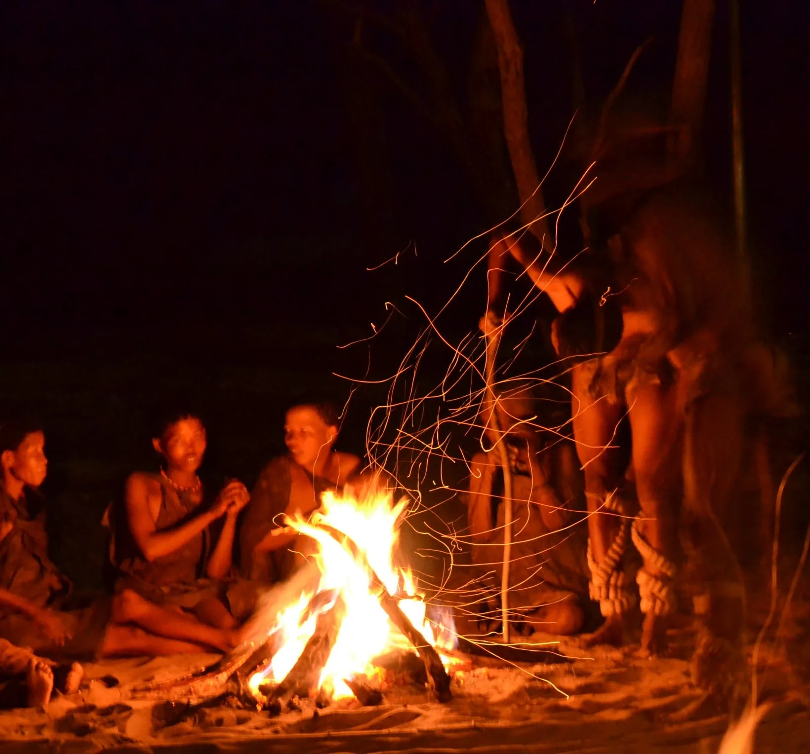 A community of Khoisan bushmen seated in a circle around a large bonfire at night, men performing traditional dances as sparks fly off the fire in the wind